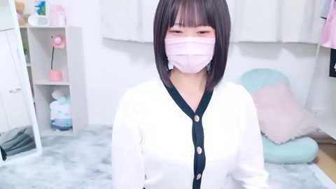 Media: Video of an Asian woman with short black hair and light skin, wearing a white cardigan, pink face mask, and standing in a pastel-colored bedroom with white walls, shelves, and a plush rug.