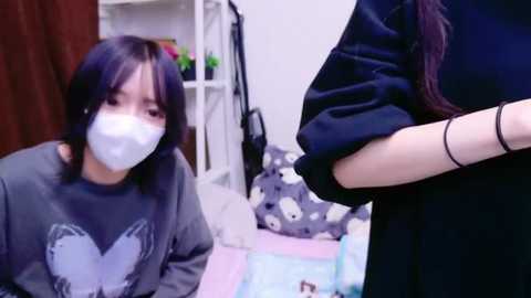 Media: Video of an Asian woman with a mask, grey shirt, and glasses kneeling on a pastel-patterned floor, facing a person in black clothing standing in the foreground, with a white shelf and toys in the background.