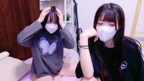 Media: Video of two young Asian women with long black hair, wearing white face masks and grey sweaters, sitting on a bed, adjusting their masks. Background includes a brown curtain and white furniture.