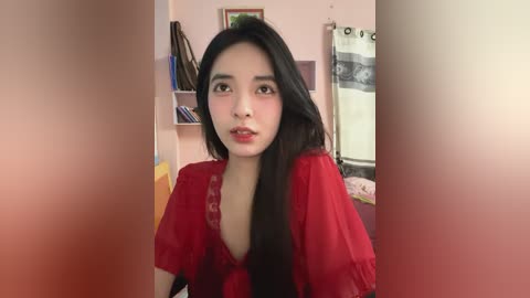 Media: Video of a young Asian woman with long black hair, wearing a red sheer blouse, seated indoors. Background features a pink wall, bookshelf, and curtains.