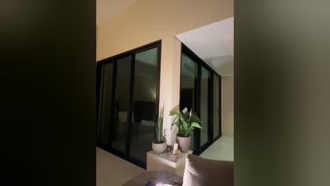 Media: Video of a modern, minimalist balcony with beige walls, large black-framed sliding glass doors, a small round table with potted plants, and a beige cushioned chair.