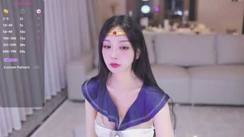 Media: A video of an East Asian woman in a Sailor Moon cosplay, with long black hair, a purple sailor collar, and a gold tiara. She stands in a modern living room with white furniture and a soft, blurred background.