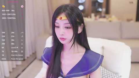 Media: Video of an East Asian woman with long black hair, wearing a Sailor Moon costume with a blue sailor collar and a red headband. She sits on a white couch in a modern living room.