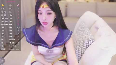 Media: Video of a young East Asian woman in a Sailor Moon cosplay, sitting on a couch with a white cushion, wearing a blue sailor collar and a yellow headband. Background shows a room with a TV screen displaying a game interface.