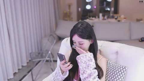 Media: Video of an Asian woman with long black hair, wearing a white sweater, using a smartphone with glasses on, sitting on a white couch in a modern, minimalistic living room with white curtains and a white dresser in the background.