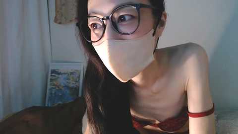 Media: Video of an Asian woman with long black hair, wearing large black glasses, a red bra, and a surgical mask. She is indoors, in a dimly lit room with a beige wall and a framed poster in the background.