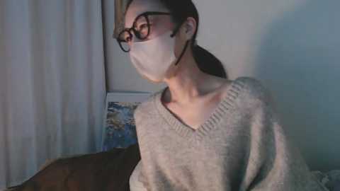 Media: Video of a woman with light skin, brown hair tied back, wearing a white mask, black-framed glasses, and a gray sweater, standing indoors by a window with white curtains and a blue painting in the background.