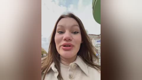 Media: Video of a young woman with light skin, brown hair, and blue eyes, wearing a beige coat, smiling with her mouth open, set against an urban background with a green building and cloudy sky.