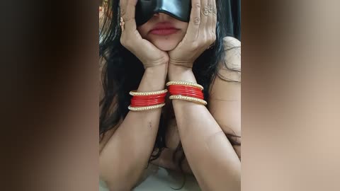 Media: A video of a woman with long black hair, wearing a black blindfold and red bangles on her wrists, her hands cupping her face.