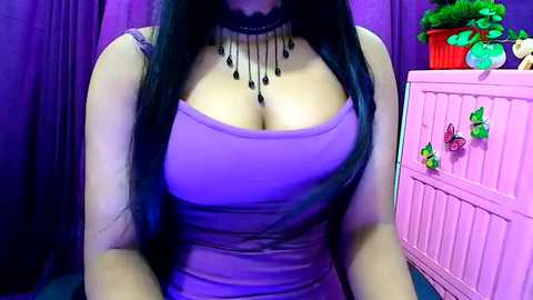 Media: Video of a woman with long black hair, wearing a low-cut, strapless purple dress that accentuates her cleavage. She has a multi-strand black necklace. The background features pink furniture with hanging decorations and a green plant.