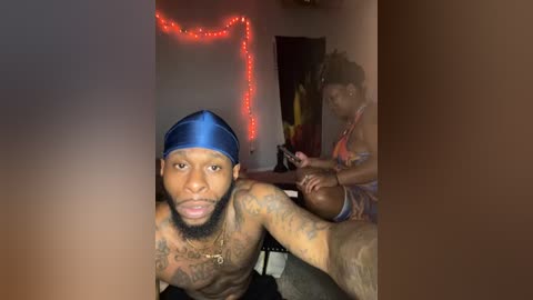 Media: Video of a shirtless, tattooed Black man with a blue headband, holding a camera in a dimly lit room with red string lights. Another Black man, wearing a colorful shirt, is seated nearby, playing with a football.