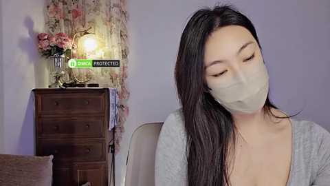 Media: Video of an Asian woman with long black hair, wearing a beige surgical mask, gray top, and seated indoors. Background features a floral-patterned curtain, wooden dresser, and a lamp.