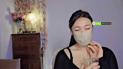 Media: Video of an Asian woman with long black hair, wearing a beige mask, drinking from a clear glass, in a cozy room with floral wallpaper, wooden dresser, and a green pillow.