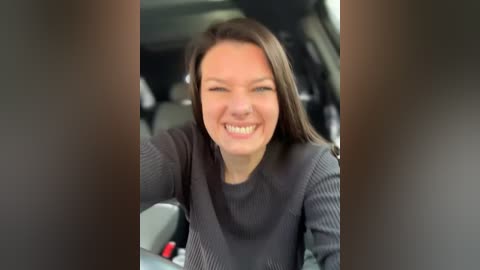 Media: Video of a smiling, fair-skinned woman with long brown hair, wearing a gray knitted sweater, sitting in the driver's seat of a car.