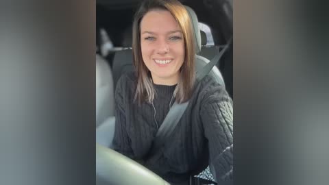 Media: Video of a smiling woman with light skin and shoulder-length brown hair, wearing a gray sweater, sitting in a car with a gray seatbelt visible.