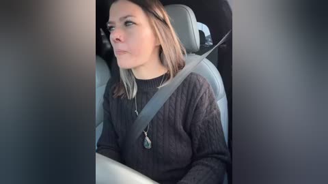 Media: Video of a woman in a car, wearing a dark knitted sweater, light skin, shoulder-length light brown hair, grey seatbelt, side profile, looking out the window.