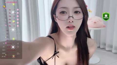 Media: A video of an Asian woman with long brown hair, glasses, and a black lace bra, taking a selfie in a bright, white room with pink decor.