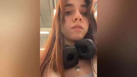 Media: Video of a young woman with long, straight, auburn hair, wearing large black headphones, and a star necklace, indoors with a ceiling light.