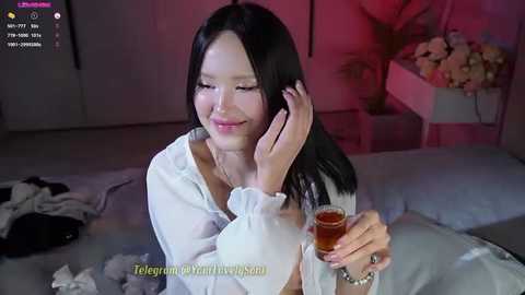 Media: Video of an East Asian woman with long black hair, wearing a white blouse, smiling while holding a glass of whiskey, in a dimly lit bedroom.