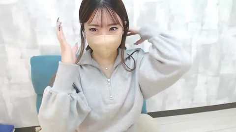 Media: Video of an East Asian woman with long black hair, wearing a light gray hoodie, surgical mask, and black nail polish, adjusting her hair in a white-tiled room.