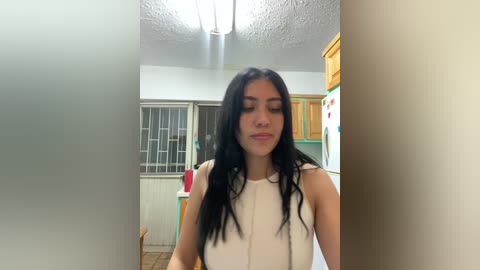 Media: Video of a young woman with long black hair, wearing a white sleeveless top, standing in a modest kitchen with wooden cabinets, a refrigerator, and a window.