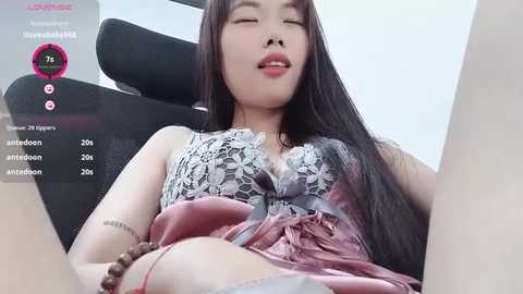 Media: A video of an East Asian woman with long black hair, wearing a lace top and pink skirt, lying on a black chair, eyes closed, in a minimalist room with a live-stream overlay.