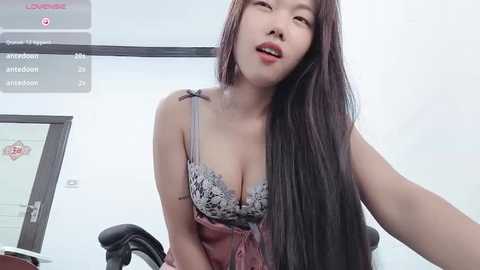 Media: Video of an East Asian woman with long black hair, fair skin, and red lipstick, wearing a pink camisole with a lace bra, seated on a black chair in a modern office.