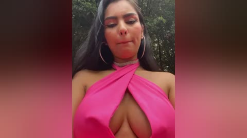 Media: Video of a woman with long black hair and large hoop earrings, wearing a bright pink halter dress with a deep neckline that reveals her cleavage, set against a blurred green background.