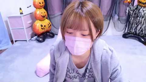 Media: Video of a young Asian girl in a gray hoodie, pink mask, kneeling on a light carpet, surrounded by Halloween decorations (jack-o'-lanterns, zebra-print bag).