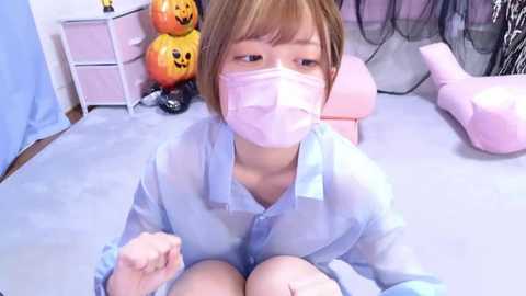 Media: Video of a young woman with short brown hair, wearing a light blue button-up shirt, and a pink face mask, sitting on a bed with a pink pillow. Background includes Halloween decorations and a pink storage unit.