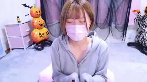 Media: Video of a young Asian woman with short brown hair, wearing a pink face mask, gray hoodie, and sitting cross-legged on a light carpet. Background features a pink wall, Halloween decorations, and a zebra-patterned rug.