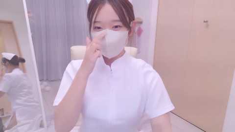 Media: Video of an Asian woman with light skin, wearing a white mask, white dress, and hair tied back, standing in a hospital room with beige walls and a gray curtain.