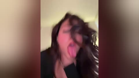 Media: A blurry video of a person with dark hair, making a silly face with tongue out, wearing a black shirt. Background is out of focus.