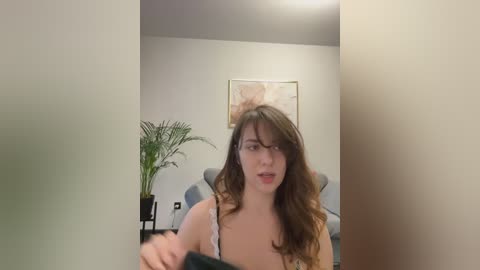 Media: Video of a young woman with long brown hair, wearing glasses, and a black lace bra, captured in a modern, minimalist living room with a beige couch, potted plant, and abstract wall art.