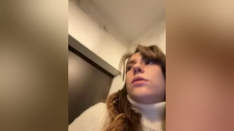 Media: Video of a woman with long, brown hair and fair skin, wearing a white turtleneck sweater, taken from a low angle, partially blurred, in a dimly lit room with beige walls.