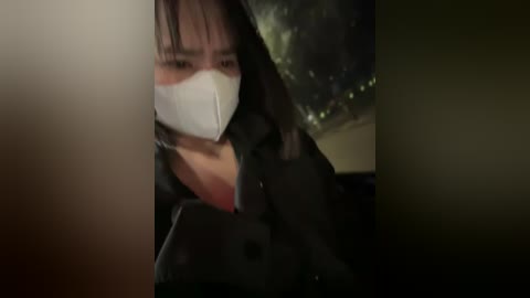 Media: A dimly lit video of a woman with a white surgical mask, black hair, and a black coat, sitting in a car, her face obscured by the camera's angle.