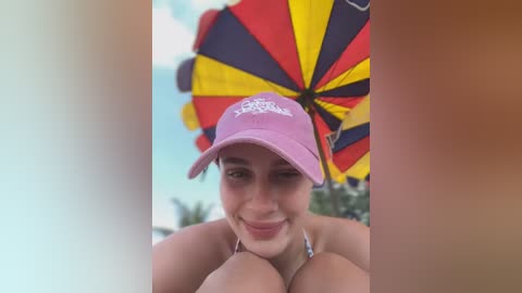 Media: Video of a smiling young woman with light skin and dark hair, wearing a pink cap and bikini, holding a colorful umbrella.