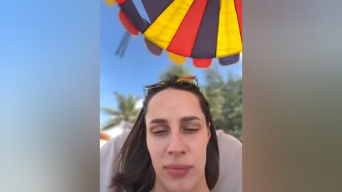 Media: A video of a woman with long dark hair, closed eyes, and sunglasses atop her head, standing in front of a colorful umbrella with yellow, red, and blue sections against a blurred beach background.