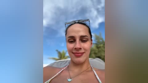 Media: Video of a woman with medium skin tone and dark hair, wearing sunglasses, a white bikini top, and a necklace, smiling against a cloudy sky and palm tree background.