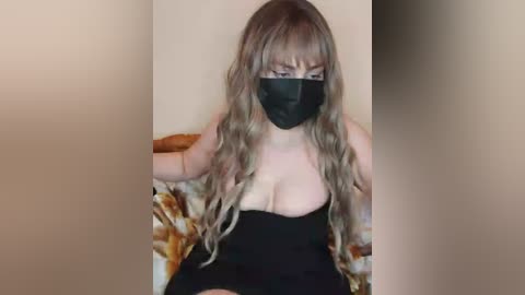 Media: Video of a light-skinned woman with long, wavy brown hair and bangs, wearing a black face mask, low-cut black top, and floral-patterned blanket. She is seated indoors, partially obscured by the camera's focus blur.