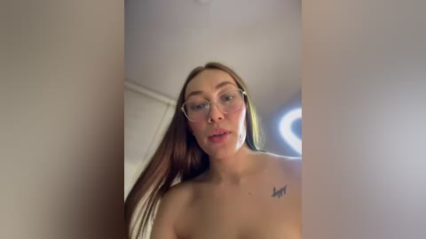 Media: A video of a topless young woman with long brown hair, wearing glasses, and a small tattoo on her right shoulder. She stands in a dimly lit room with a ceiling light.