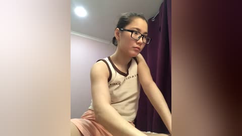 Media: A young Caucasian woman with glasses, light skin, and brown hair tied back, wearing a white tank top and pink shorts, sits on a bed in a dimly lit room with purple curtains.