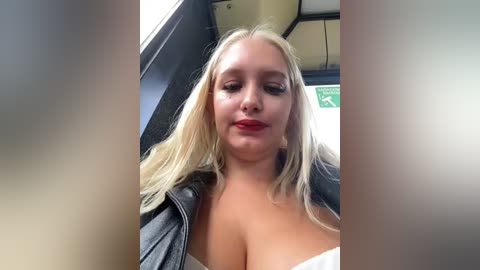 Media: A video of a blonde woman with fair skin, wearing a white bra and a black leather jacket, sitting in a car with a blurred background.