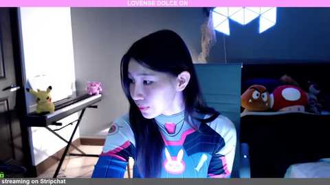 Media: Video of a young woman in a white and pink Overwatch-themed jacket, sitting in a dimly lit room with a Pikachu plush toy, a Mario plush, and a computer.