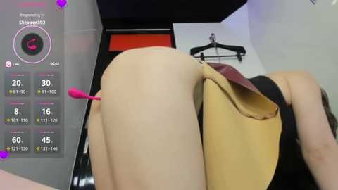 Media: Video of a woman bending over, her beige dress pulled up to reveal her buttocks, with a pink vibrator inserted. Background shows a modern, dimly lit room with a black hanger.