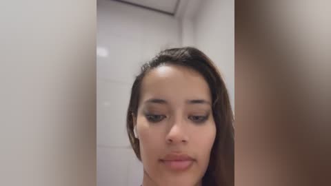 Media: Video of a young woman with medium brown skin and long, straight black hair, wearing dark eyeliner and subtle makeup. She stands in a narrow hallway with white walls and brown trim.