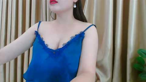 Media: Video of a light-skinned woman with long brown hair, wearing a deep blue, lace-trimmed camisole. She has red lipstick and pearl earrings. Background features gold curtains and a green potted plant.