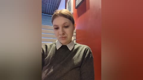 Media: Video of a young woman with light skin and brown hair, wearing a gray sweater over a white collared shirt, standing in a hallway with orange walls and a partially visible window with closed blinds.