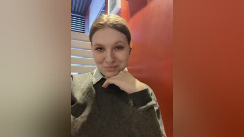 Media: Video of a young Caucasian woman with light skin and brown hair, wearing a gray sweater, standing indoors with a red wall and white blinds in the background.