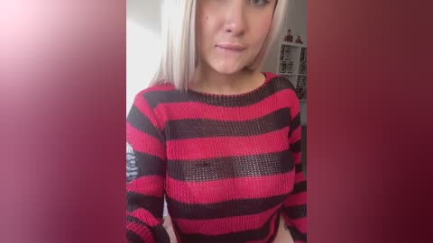 Media: Video of a young Caucasian woman with straight, shoulder-length platinum blonde hair, wearing a red and black striped knit sweater. She has a neutral expression and is indoors, with a blurred background.